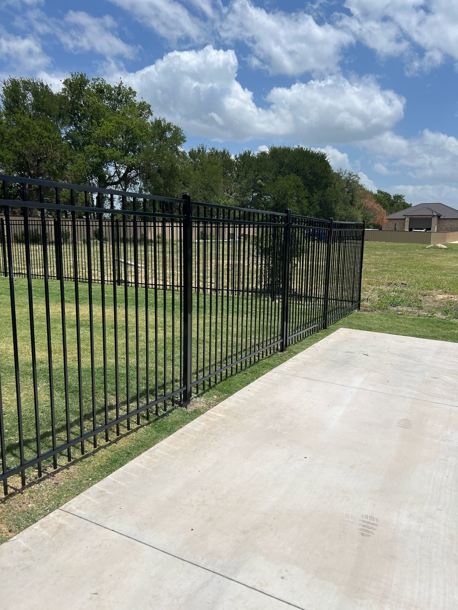 Outdoor residential backyard with a black metal fence enclosing a grassy area, bordered by trees and dense greenery; a concrete patio or foundation slab is visible in the foreground under clear daylight.